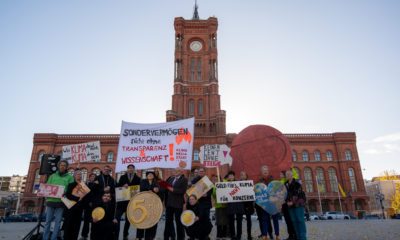 Foto einer Gruppe von Menschen, die vor dem Roten Rathaus stehen. Es sieht aus wie eine Demonstration, denn viele der Menschen halten Schilder hoch. Das größte ist als Art Münze gestaltet, darauf steht: 5 Mrd. Euro. Auf einem Transparent steht: "Sondervermögen nicht ohne Transparenz und Wissenschaft"