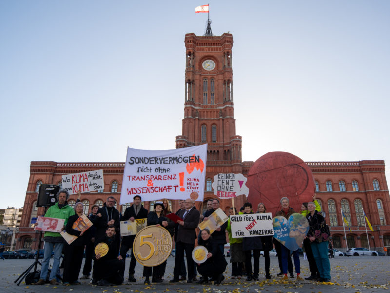 Foto einer Gruppe von Menschen, die vor dem Roten Rathaus stehen. Es sieht aus wie eine Demonstration, denn viele der Menschen halten Schilder hoch. Das größte ist als Art Münze gestaltet, darauf steht: 5 Mrd. Euro. Auf einem Transparent steht: "Sondervermögen nicht ohne Transparenz und Wissenschaft"