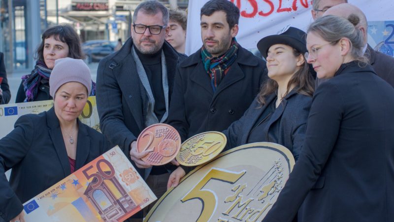 Bild einer Demonstration, mehrere Menschen stehen zusammen und halten eine große Münze auf Pappe hoch, auf der steht: 5 Mrd. Euro