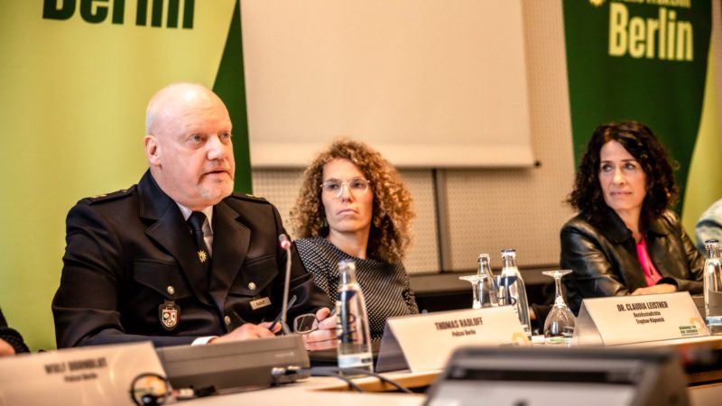 Ein Polizist in Uniform (Thomas Radloff) sitzt in Uniform an einem Tisch neben Claudia Leistner und Bettina Jarasch. Er spricht mit ernstem Blick.