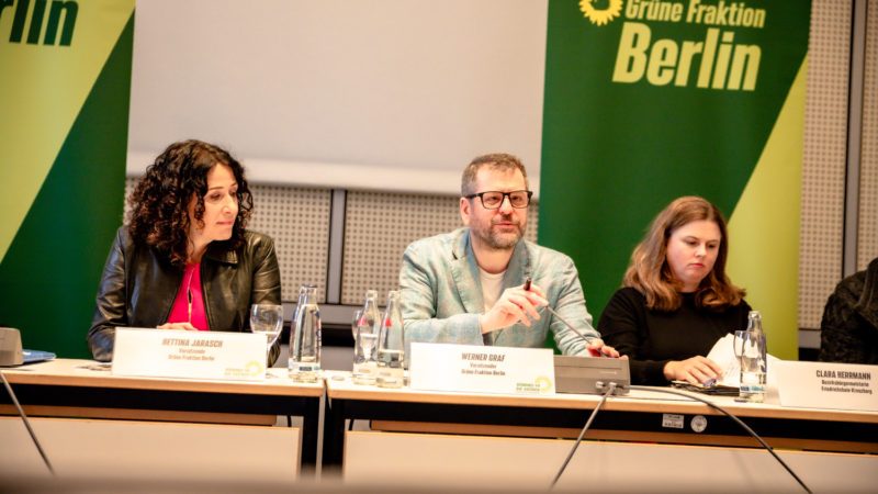 Bettina Jarasch, Werner Graf und Clara Herrmann sitzen nebeneinander an einem Tisch. Im Hintergrund zwei grüne Banner.