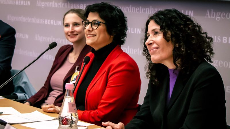 Drei Frauen sitzen auf einem Podium nebeneinander. Die beiden rechten Frauen sind Bettina Jarasch und Gollaleh Ahmadi. Sie schauen beide nach vorn und lächeln.