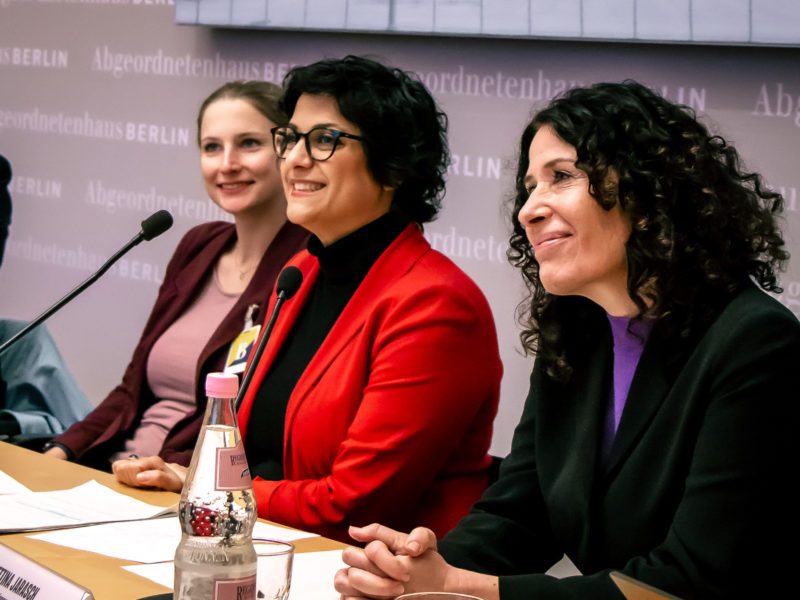 Drei Frauen sitzen auf einem Podium nebeneinander. Die beiden rechten Frauen sind Bettina Jarasch und Gollaleh Ahmadi. Sie schauen beide nach vorn und lächeln.