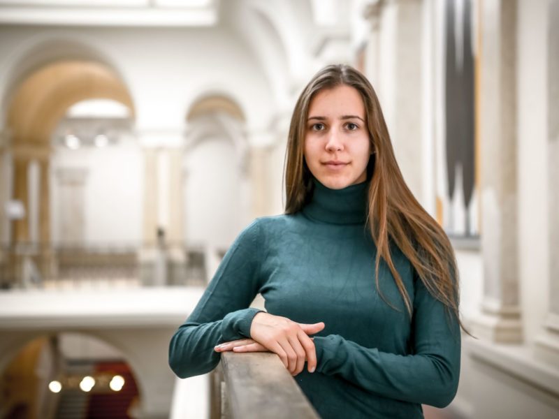 Klara Schedlich steht auf einer Galerie im Abgeordnetenhaus und stützt eine Hand auf dem Geländer auf. Ihr Blick ist ernst. Sie ist eine junge Frau mit langen braunen Haaren.