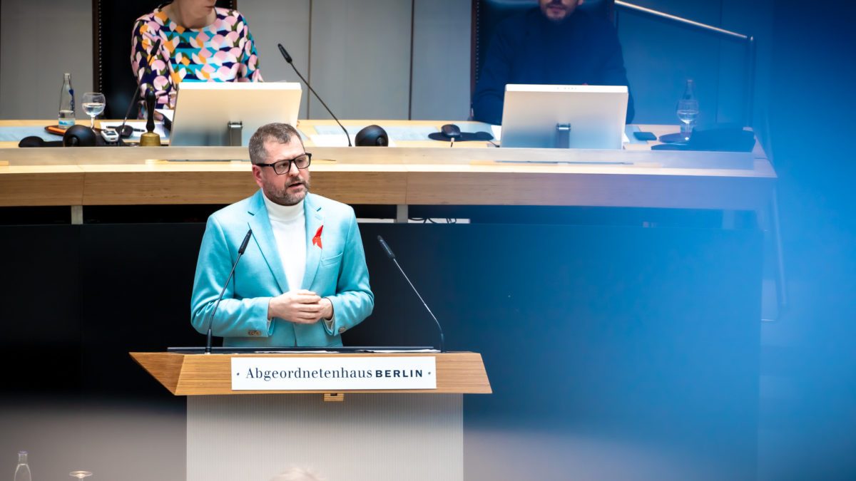 Werner Graf steht am Redepult des Abgeordnetenhauses. Er trägt einen hellblauen Blazer und eine rote AIDS-Schleife. Sein Blick ist ernst.