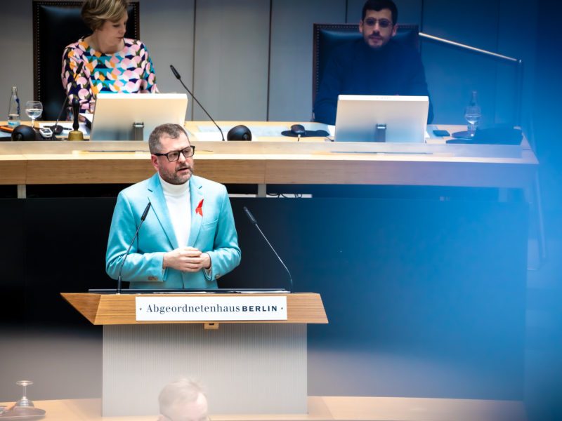 Werner Graf steht am Redepult des Abgeordnetenhauses. Er trägt einen hellblauen Blazer und eine rote AIDS-Schleife. Sein Blick ist ernst.