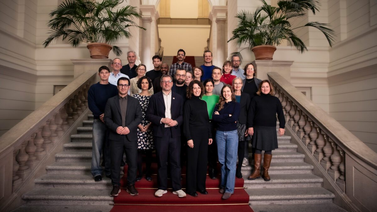 Gruppenfoto der Grünen Fraktion Berlin nach der konstituierenden Sitzung. Etwa 25 Abgeordnete stehen in mehreren Reihen gestaffelt auf einer hellen Steintreppe mit rotem Teppich und steinernen Geländern im Abgeordnetenhaus von Berlin. Die meisten lächeln in die Kamera, viele tragen dunkle Kleidung, einzelne setzen Farbakzente mit einem grünen und einem roten Pullover. Links und rechts oben stehen große Palmen in Terrakottatöpfen, im Hintergrund führt die Treppe zu einem hell erleuchteten Torbogen.